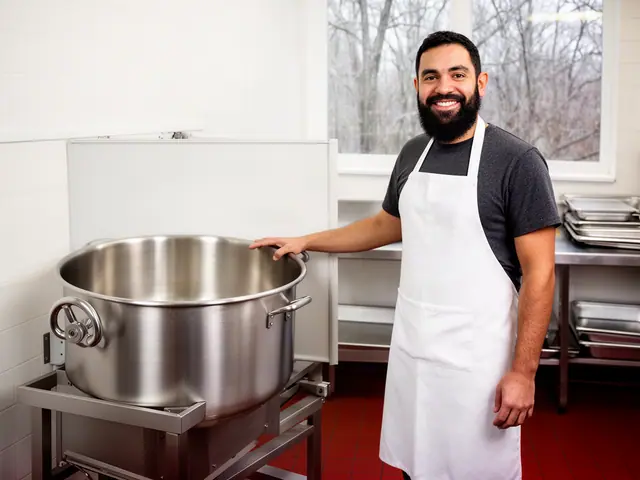 A smiling cook in a white apron beside a new stainless-steel tilting kettle in a commercial kitchen.